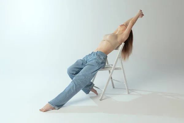 Tender bela menina posando em roupas íntimas e jeans na cadeira isolada sobre fundo estúdio cinza. Auto-amor — Fotografia de Stock