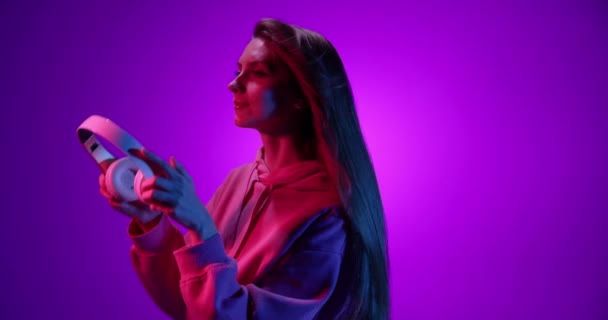Šťastně se usmívající mladá dívka poslouchající hudbu ve sluchátkách a tančící na purpurovém pozadí v neonovém světle, filtr. 4K. Emoce, reklamy, kultura, koncepce mládeže — Stock video