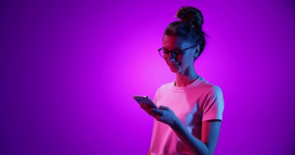 Emotional young girl in eyewear using phone, watching funny video isolated on purple background in neon light, filter. 4K. Emotions, ads, culture, youth concept — Stock Video