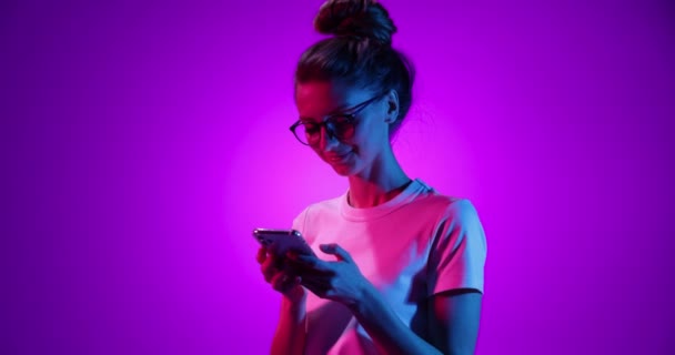 Щаслива молода дівчина в окулярах за допомогою телефону, дивиться смішне відео ізольоване на фіолетовому фоні в неоновому світлі, фільтр. 4K. Емоції, реклама, культура, концепція молоді — стокове відео