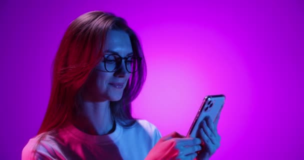 Hermosa chica joven en gafas usando gadget, desplazamiento alimentación aislada sobre fondo púrpura en luz de neón, filtro. 4K. Emociones, anuncios, cultura, concepto de juventud — Vídeo de stock
