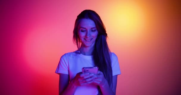 Spännande ung flicka i glasögon med hjälp av telefon, titta på rolig video isolerad på röd-gul bakgrund i neonljus, filter. 4K. Känslor, annonser, kultur, ungdomskoncept — Stockvideo