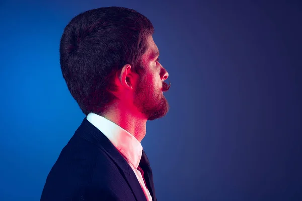 Closeup. Profile view of young stylish bearded man in business suit posing isolated on dark blue studio background. Concept of emotions, style, sales, ad, fashion and beauty — Stock Photo, Image