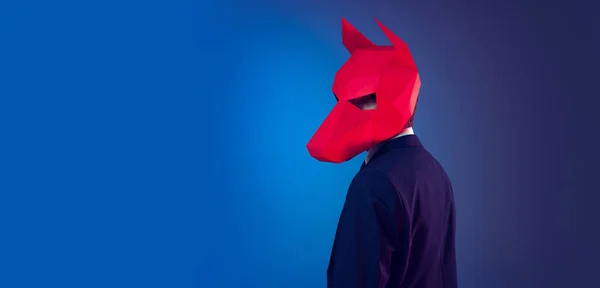 Kreativní portrét mladého stylového muže v obleku s červenou lepenkovou maskou na hlavě izolovaný na tmavomodrém pozadí studia. Koncept umění, módy, divadla — Stock fotografie