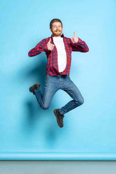 Emotional young red-headed man in white t-shirt and plaid shirt jumping isolated on blue background. Concept of art, fashion, emotions, aspiration