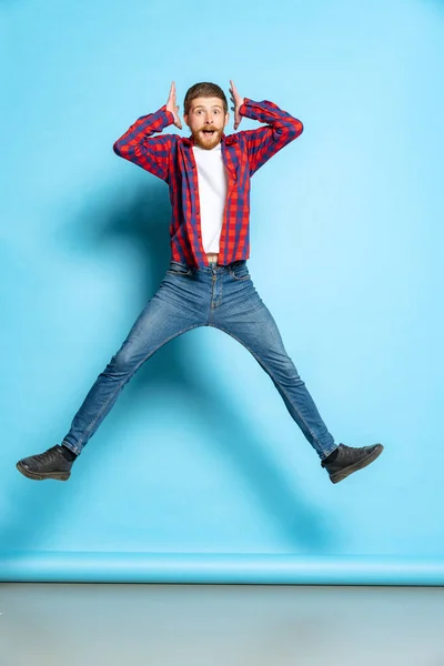 Emotional young red-headed man in white t-shirt and plaid shirt jumping isolated on blue background. Concept of art, fashion, emotions, aspiration
