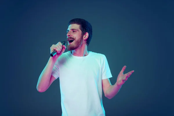 Hombre guapo en camiseta blanca cantando al micrófono aislado sobre fondo de estudio azul oscuro, concepto de emociones humanas, expresión facial, ventas, anuncio, moda y belleza — Foto de Stock