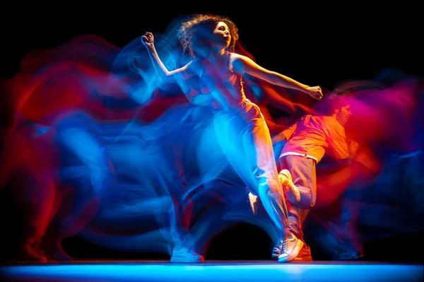 Portrait dynamique d'homme et de fille élégants dansant hip-hop dans des vêtements de style sportif sur fond sombre à la salle de danse dans la lumière au néon mixte. Culture des jeunes, breakdance, mouvement — Photo