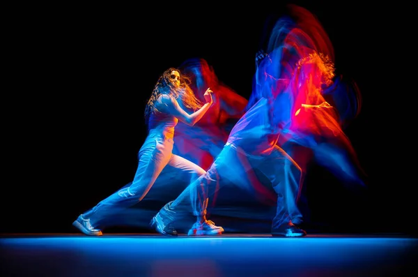 Динамичный портрет стильного мужчины и девушки, танцующих хип-хоп в спортивной одежде на тёмном фоне в танцевальном зале в смешанном неоновом свете. Молодежная культура, брейк-данс, движение — стоковое фото