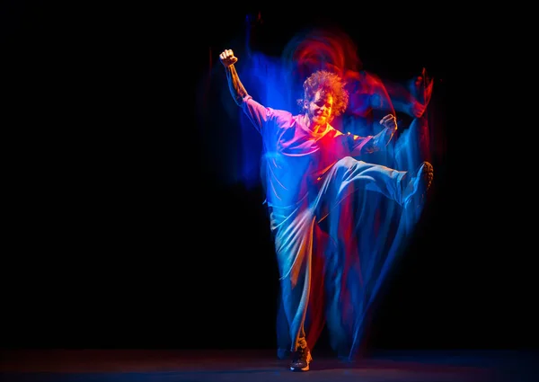 Стильный мужчина танцует хип-хоп в спортивной одежде, изолированной на тёмном фоне в танцевальном зале в смешанном неоновом свете. Молодежная культура, хип-хоп, движение — стоковое фото