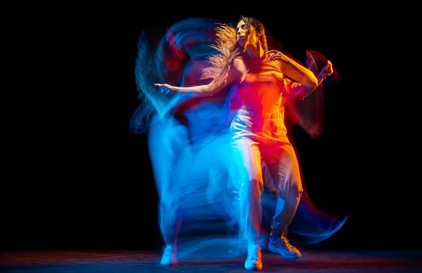 Excited man and woman dancing hip-hop in sportive style clothes on dark background at dance hall in mixed neon light. Youth culture, hip-hop, movement