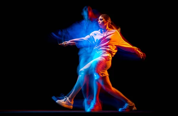 Young woman dancing hip-hop in sportive style clothes isolated on dark background at dance hall in mixed neon light. Youth culture, hip-hop, movement