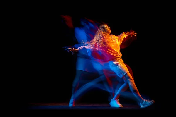 Молодая женщина танцует хип-хоп в спортивной одежде, изолированной на тёмном фоне в танцевальном зале в смешанном неоновом свете. Молодежная культура, хип-хоп, движение — стоковое фото