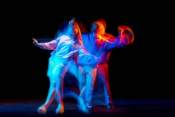 Dynamisch portret van stijlvolle man en meisje dansend hip-hop in sportieve stijl kleding op donkere achtergrond in danszaal in gemengd neonlicht. Jeugdcultuur, breakdance, beweging — Stockfoto