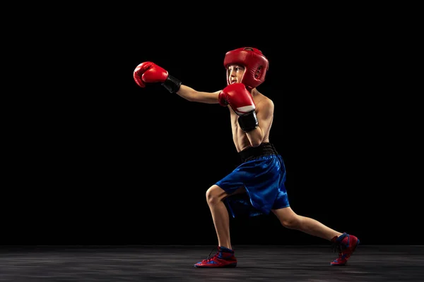 Dynamiskt porträtt av sportig pojke, unge i boxerhandskar och shorts öva isolerad på mörk bakgrund. Begreppet idrott, rörelse, studier, prestationer, aktiv livsstil. — Stockfoto