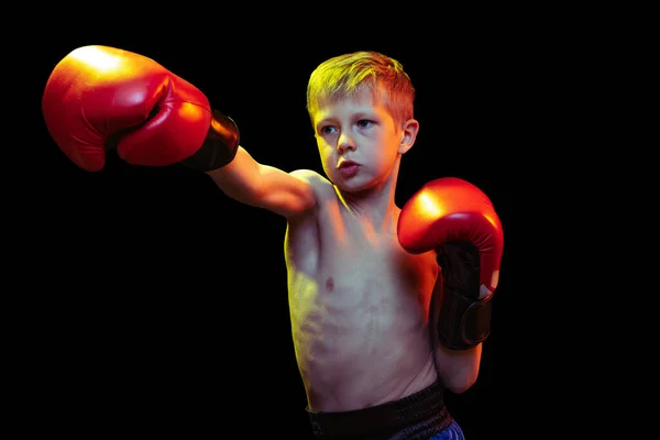 Pequeño chico deportivo, boxeador principiante con guantes de boxeador rojo y pantalones cortos entrenando aislado sobre fondo oscuro. Concepto de deporte, movimiento, estudio, logros, estilo de vida activo. —  Fotos de Stock