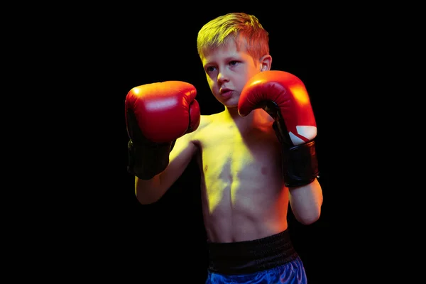 Little sportive boy, beginner boxer in red boxer gloves and shorts training isolated on dark background. Concept of sport, movement, studying, achievements, active lifestyle. — Stock Photo, Image