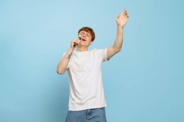 Şarkıcı. Stüdyoda, beyaz tişörtlü, mavi stüdyo arka planında yalnız poz veren genç, kızıl saçlı bir adam var. Gençlik kavramı, moda, duygular, yüz ifadesi. İlan için alanı kopyala