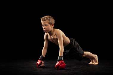 Şınav. Anaokulu çocuğu, spor çekimlerinde tatlı bir çocuk ve tek başına siyah arka planda antrenman yapıyor. Annemin hareket, hareket, spor, meydan okuma kavramının acemi dövüşçüsü. İlan için alanı kopyala