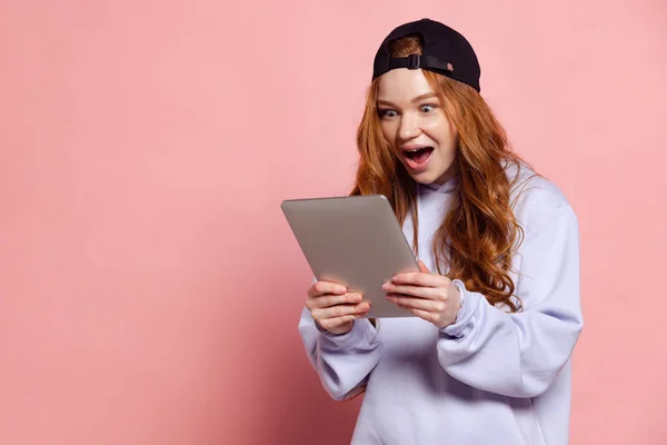 Wow, surprise. Young redhead girl, student in hoodie and cap using tablet isolated on pink studio background. Fashion, emotions, facial expression concept. Trendy colors. Youth culture, sales