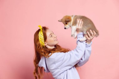 Yeni bir arkadaş. Uzun kıvırcık saçlı neşeli kızıl saçlı genç kız pembe arka planda küçük şirin bir köpek yavrusu tutuyor. İlgi, destek ve sevgi. Gençlik, güzellik, hayat kavramı. Uzayı kopyala