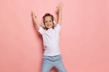 Dans etmek ve müzik dinlemek. Küçük şirin bir kızın stüdyo fotoğrafı, beyaz tişörtlü bir çocuk pembe arka planda yalnız poz veriyor. Çocuk duyguları, moda, güzellik, okul ve reklam kavramı.