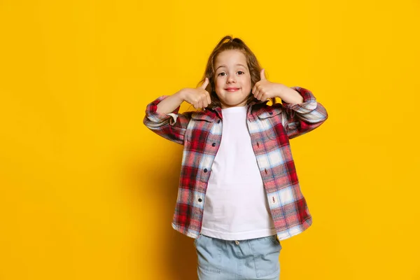 Thumbs up, nice sign. Happy little girl wearing warm plaid shirt isolated on bright yellow background. Concept of children emotions, fashion, beauty, school and ad concept. Kids fashion, street style