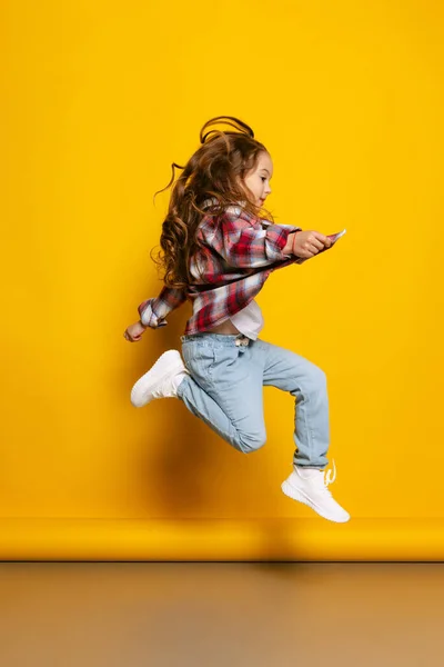 Jumping. Happy little girl, kid wearing warm plaid shirt having fun isolated on bright yellow background. Concept of children emotions, fashion, beauty, school and ad concept. Kids fashion