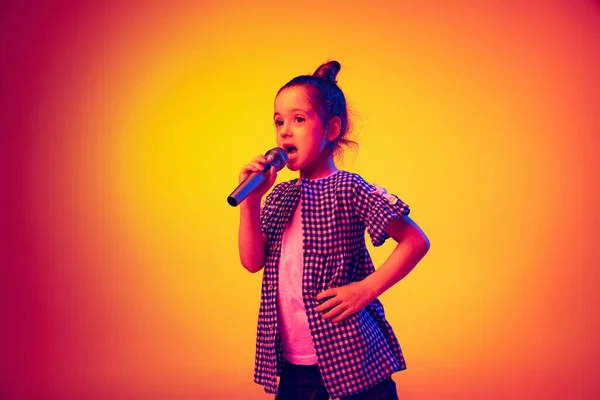Singing with microphone. Emotional cute little girl, kid posing isolated on yellow red color background in neon light. Concept of children emotions, fashion, beauty, school and ad concept.
