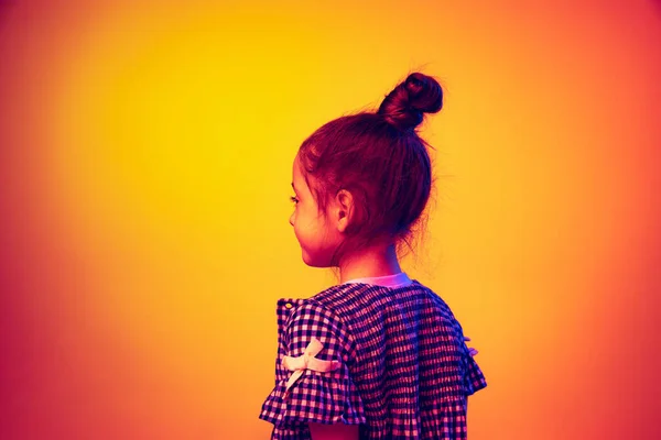 Back view of little girl, kid posing isolated on yellow red color background in neon light. Concept of children emotions, fashion, beauty, school and ad concept.