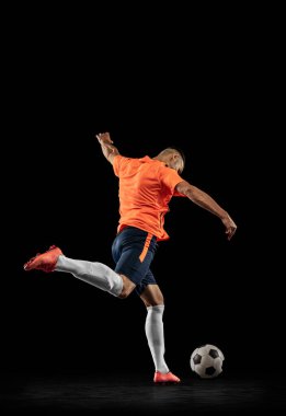 Karanlık arka planda izole edilmiş, hareketli profesyonel erkek futbol oyuncusunun dinamik portresi. Spor anlayışı, hedefler, rekabet, hobi, reklam. Turuncu-mavi futbol takımı giyen sporcular