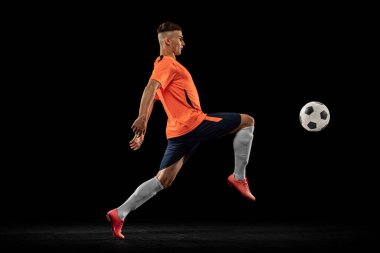 Karanlık arka planda izole edilmiş, hareketli profesyonel erkek futbol oyuncusunun dinamik portresi. Spor anlayışı, hedefler, rekabet, hobi, reklam. Turuncu-mavi futbol takımı giyen sporcular