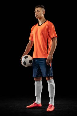 Turuncu-mavi futbol takımındaki genç erkek futbolcunun koyu arka planda topla poz verişi. Spor anlayışı, hedefler, rekabet, erkek hobisi, meslekler. Uzayı kopyala