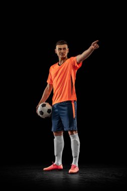 Full-length portrait of young male soccer player in orange-blue football kit posing with ball isolated on dark background. Concept of sport, goals, competition, male hobby, occupations. Copy space for clipart