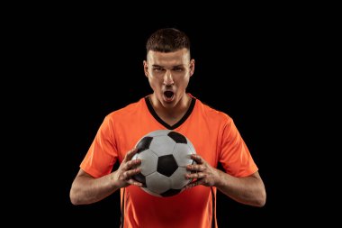 Winner. Front camera view of young male soccer player in orange-blue football kit posing with ball isolated on dark background. Concept of sport, goals, competition, occupations. Copy space for ad clipart