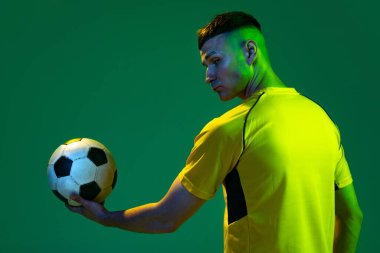 Profesyonel erkek futbolcunun topu tuttuğu yarı uzunlukta top yeşil arka planda neon ışıkta izole edilmiş. Spor anlayışı, hedefler, rekabet, oyun, başarılar. Sarı futbol takımındaki adam.