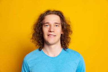 Kameraya bakıyorum. Sarı arka planda uzun kıvırcık saçlı, yakışıklı genç bir adam. Duygusal, genç yüz. Erkek yarı boy portresi. İnsani duygular, yüz ifadesi kavramı. Moda renkleri