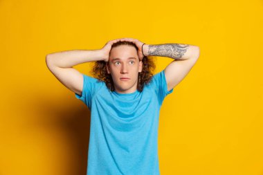 Şaşkınlık. Sarı arka planda izole edilmiş uzun kıvırcık saçlı, yakışıklı genç bir adam. Duygusal, genç yüz. Erkek yarı boy portresi. İnsani duygular, yüz ifadesi kavramı. Moda renkleri