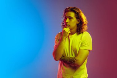 Düşünüyorum. Uzun kıvırcık saçlı, çekici genç bir adam. Gradyan mavi-pembe arka planda tek başına poz veriyor. Güzellik, moda, gençlik kültürü ve duyguları kavramı. Yarım boy erkek portresi.