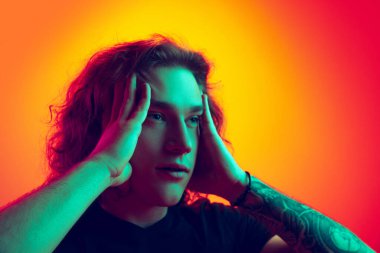 Şok oldum. Şok olmuş, uzun kıvırcık saçlı genç adam, neon renkli turuncu-pembe arka planda tek başına poz veriyor. Güzellik, moda, gençlik kültürü ve duyguları kavramı. Yakından erkek portresi.