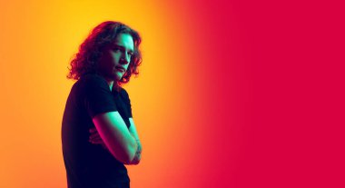 Düşünüyorum. Uzun kıvırcık saçlı, parlak sarı-mor arka planda izole edilmiş genç, ciddi bir adam. Güzellik, moda, gençlik kültürü ve duyguları kavramı. Yarım boy erkek portresi.