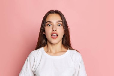 Beyaz tişörtlü genç bir kız pembe arka planda geniş gözlü kameraya bakıyor. Güzellik, sanat, moda, gençlik, satış ve reklamlar. Uzun siyah saçlı bir manken..