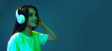 Uzun, dağınık saçlı, mavi arka planda neon ışıkta izole edilmiş müzik dinleyen çekici bir genç kız portresi. Güzellik, sanat, moda, gençlik, satış ve reklamlar. Öğrenci heyecanlı görünüyor.