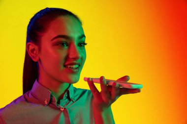 Neon ışıklarıyla izole edilmiş sarı arkaplanda izole edilmiş telefonu kullanan gülümseyen genç kızın yakın plan portresi. Duygular, yüz ifadesi, gençlik, arzu kavramı. İlan için alanı kopyala
