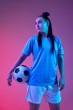 Portrait of beautiful girl, professional female soccer player with football ball isolated on purple background in neon. Concept of sport, fitness. Young spotive girl in black and white football kit clipart