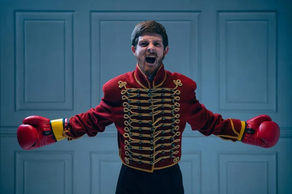 Winner emotions. Excited young man like medieval hussar wearing boxing gloves shouting isolated on dark blue background. Retro style, hobby, comparison of eras concept.