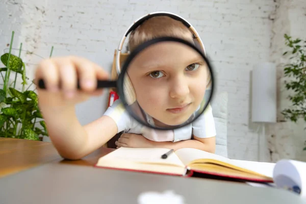 Looking through a magnifying glass. School age kid sitting at home and doing homework at home interior, indoors. Online education, childhood, people, remote learning and school concept. Looks serious