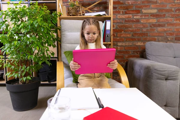 Remote learning. One beautiful little girl, school age kid studying at home using tablet. Model wearing eyeglasses. Online education, childhood, family, homework and school concept.