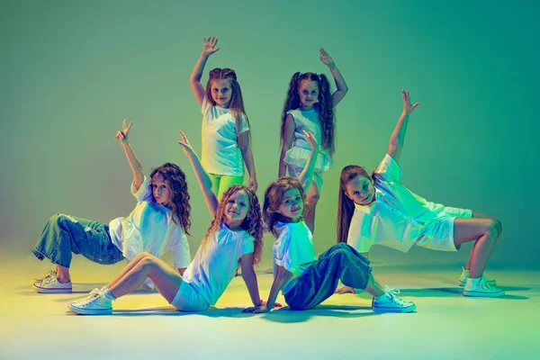 Future contemp dancers. Group of children, little girls in sportive casual style clothes dancing in choreography class isolated on green background in neon light. Concept of music, fashion, art