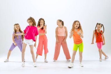 Modern koreografi dersi. Beyaz stüdyo arka planında izole halde dans eden parlak renkli elbiseli, mutlu ve aktif küçük kızlardan oluşan bir dans grubu. Müzik, moda, sanat, çocukluk, hobi kavramı.